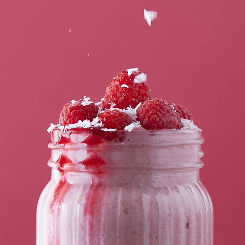 Gros plan sur la partie supérieure d'un bocal de smoothie framboises litchis saupoudré de noix de coco râpée.