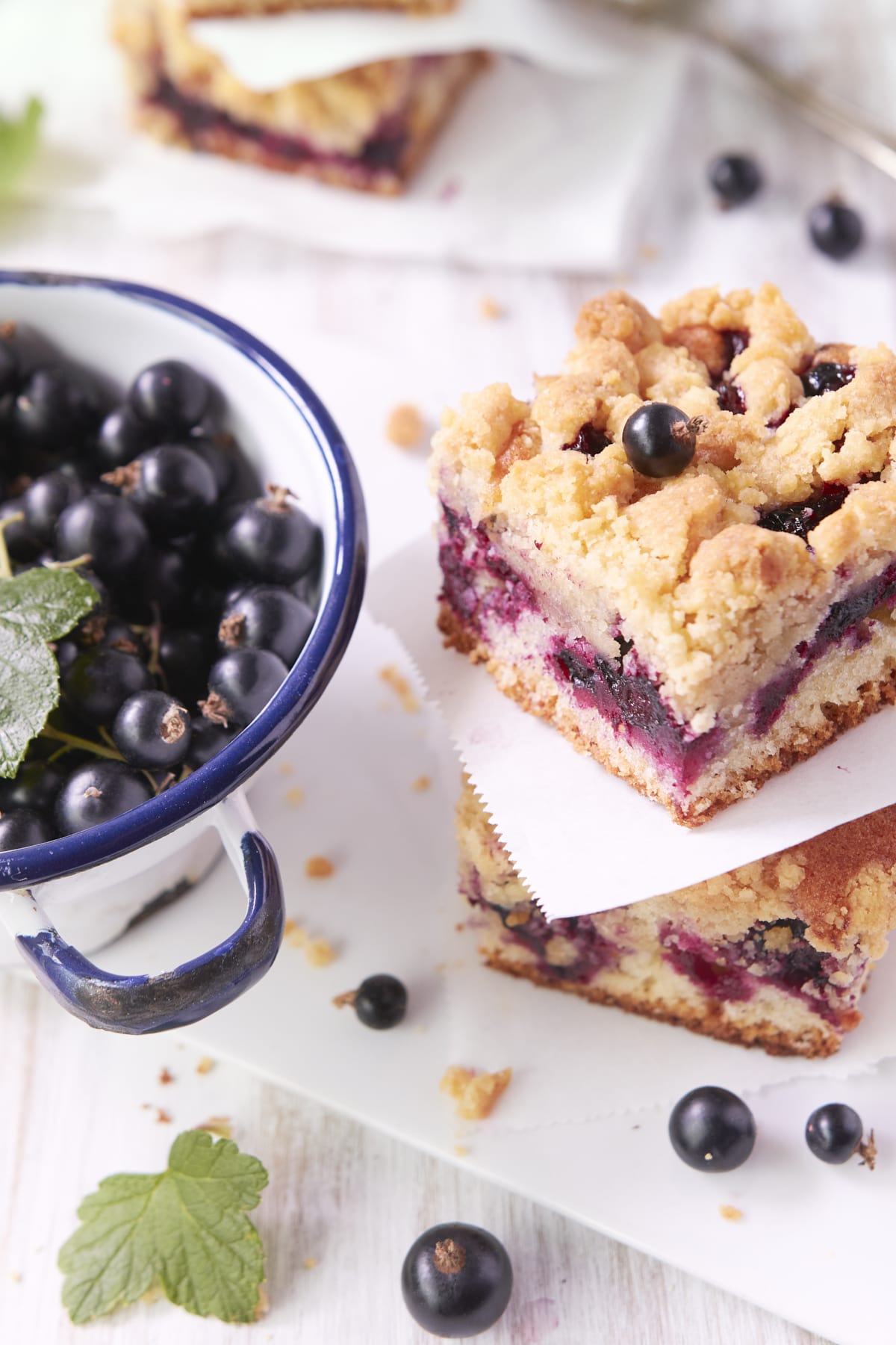 Des parts de crumb cake à côté d'un récipient en émail blanc contenant du cassis.