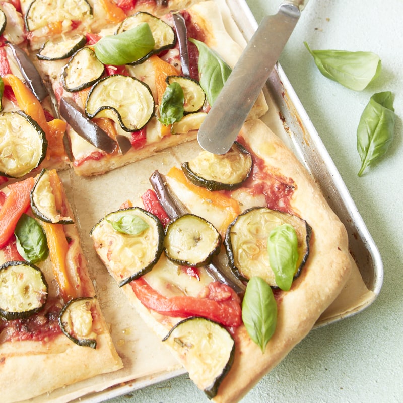Gros plan sur une pizza aux légumes grillés, découpée en parts et présentée sur une plaque de cuisson recouverte de papier sulfurisé.