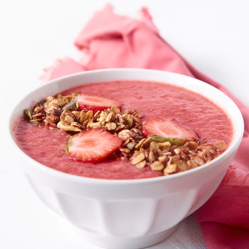 Gros plan de face sur un smoothie banane fraises garni de granola et de fraises, accompagné d'une serviette de table en tissu de la même couleur que le smoothie.