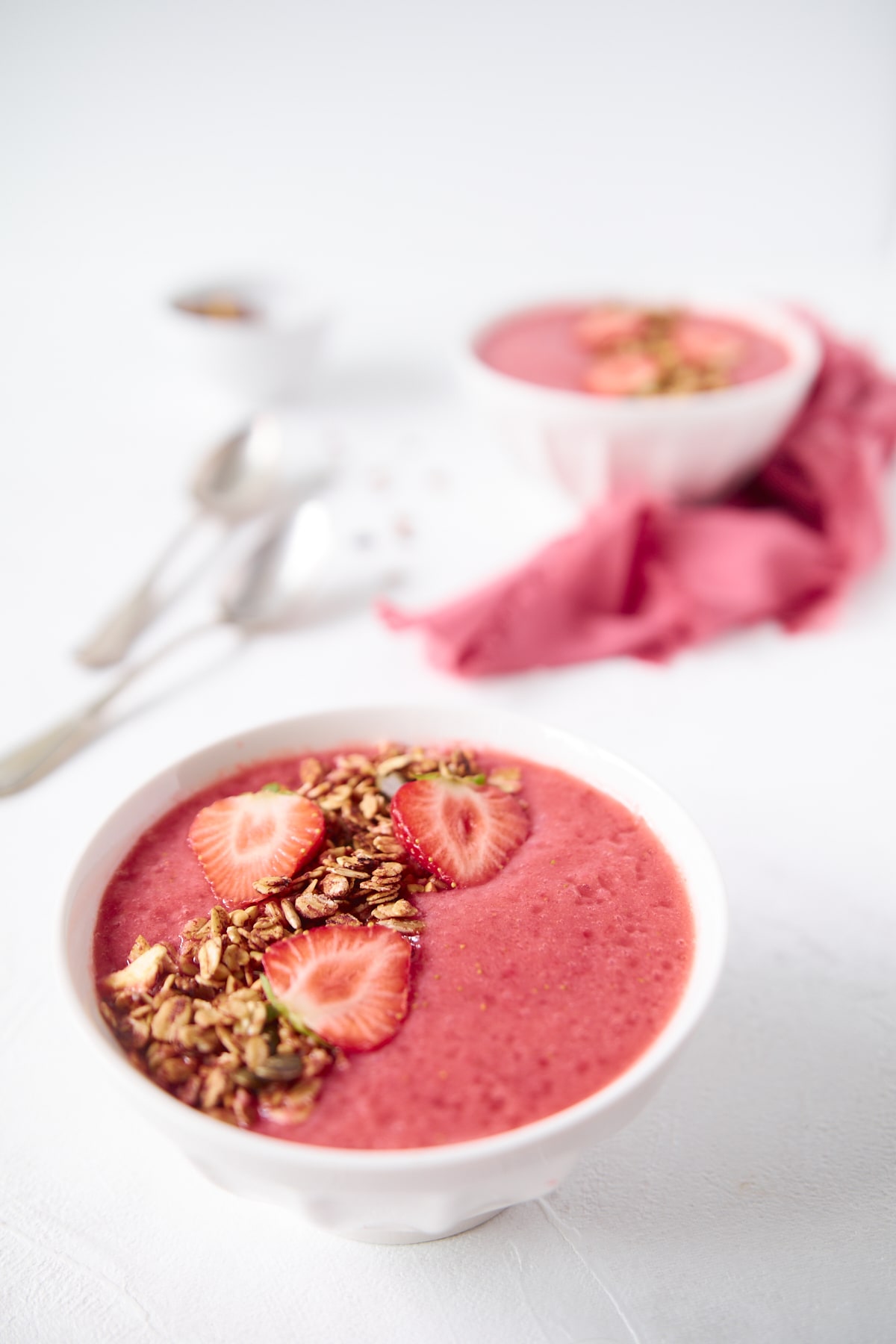 Deux bols de smoothie banane fraises décorés de granola, accompagnés de cuillères et de serviettes de tables roses.