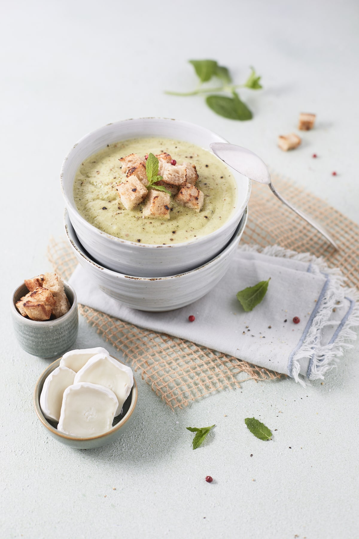 Un bol de soupe froide de courgettes à la menthe garni de croûtons et accompagné de petites tranches de fromage de chèvre.