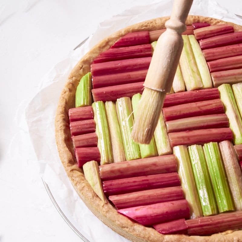 Gros plan sur une tarte géométrique à la rhubarbe en train d'être badigeonnée de sirop.