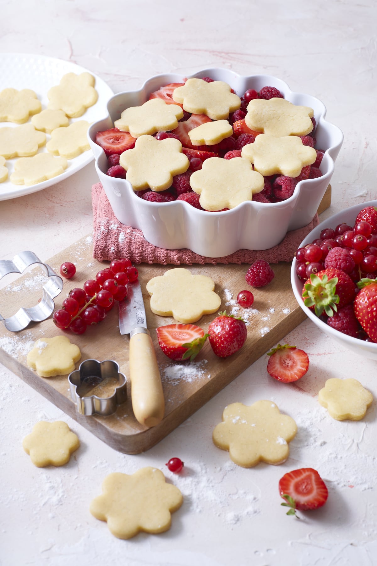 Cobbler au fruits rouges prêt à être enfourné avec, à côté du plat, des fraises, des groseilles, des framboises ainsi que des fleurs en pâte à cobbler.