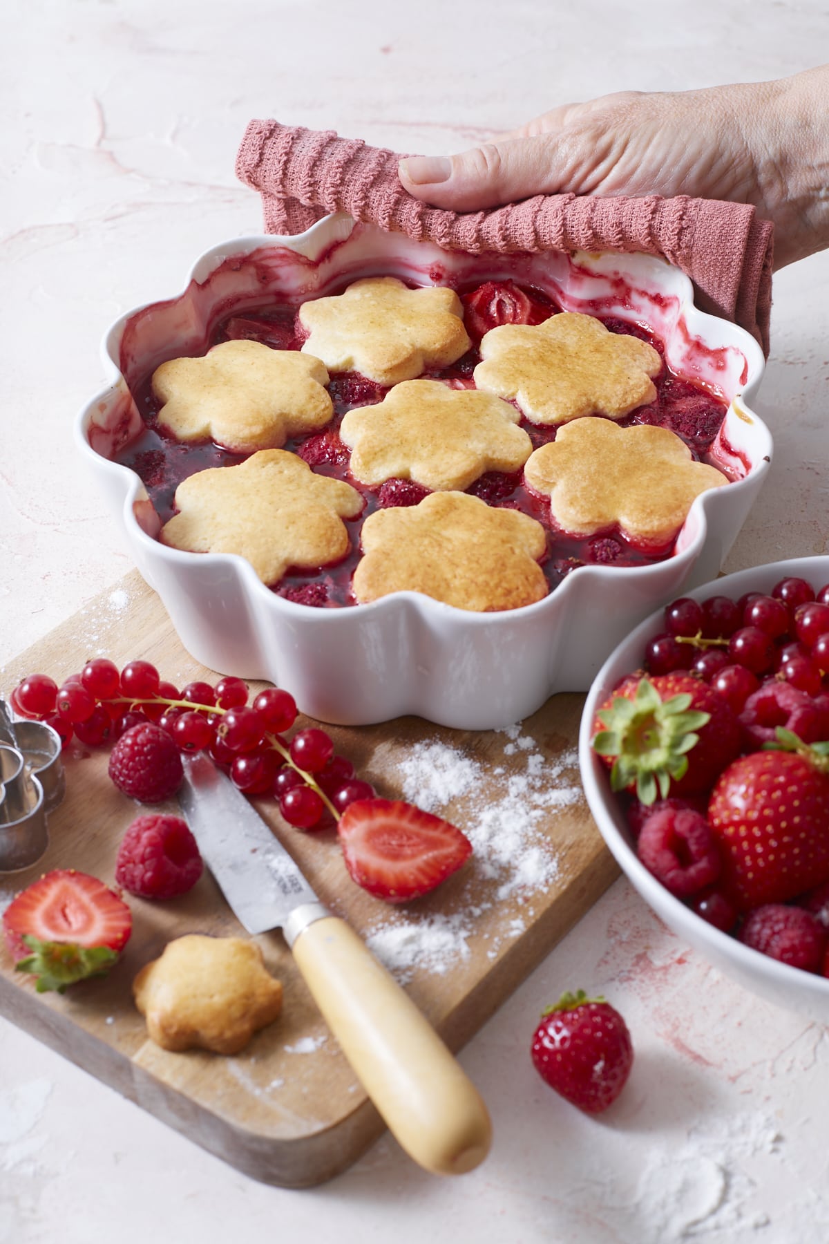 Plat de cobbler aux fruits rouges fraîchement sorti du four, tenu avec un torchon rose et entouré de fraises, framboises et grappes de groseilles rouges.