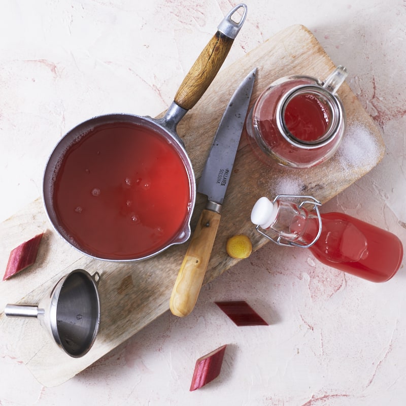 Sur une planche, une casserole et deux bouteilles de sirop de rhubarbe au gingembre.