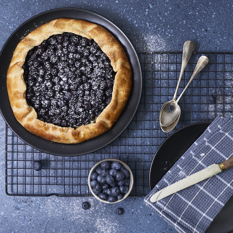 Gros plan en flatlay sur une tarte rustique aux myrtilles saupoudrée de sucre glace, présentée sur une grille.