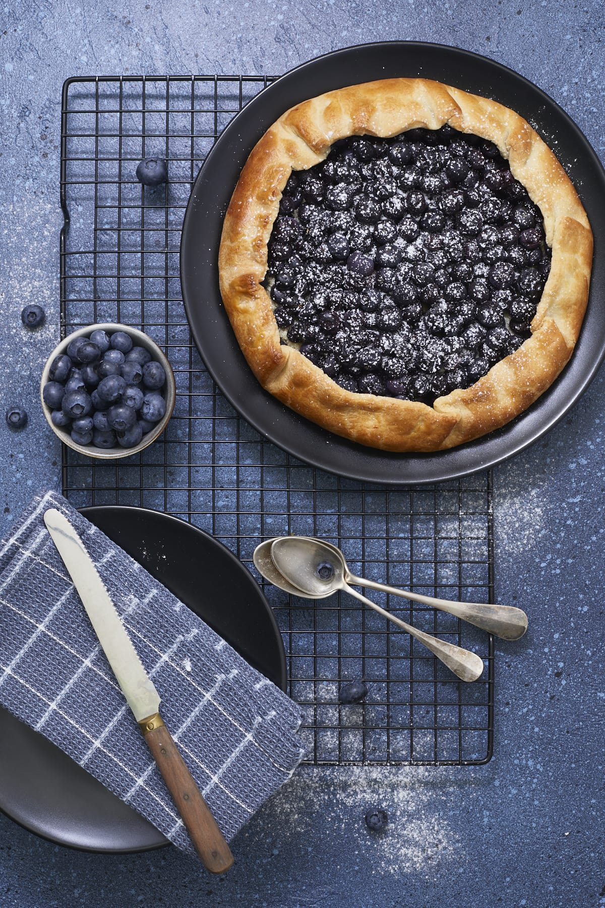 Tarte rustique aux myrtilles saupoudrée de sucre glace, présentée sur une grille avec des assiettes à dessert.