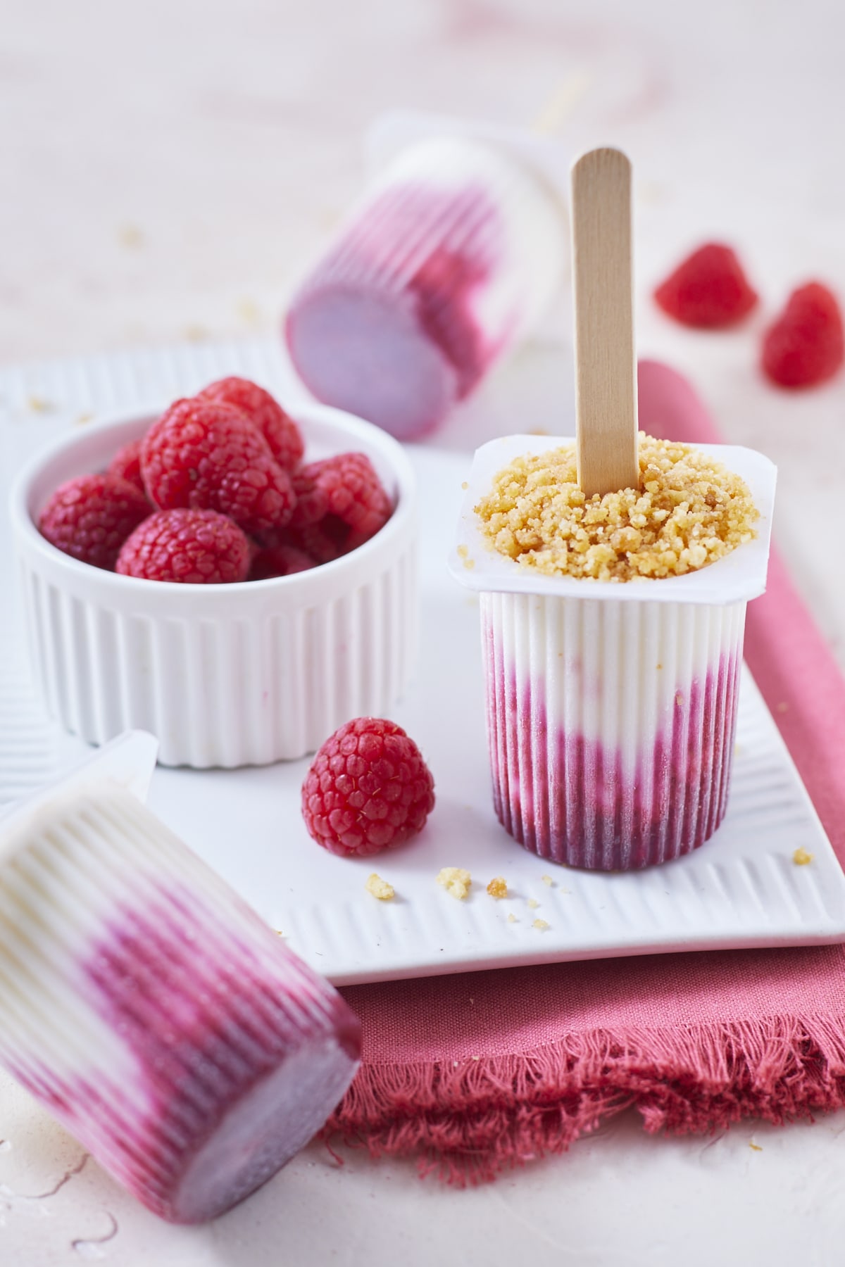 Trois petits Suisses glacés, façon cheesecake aux framboises sur un petit plat de céramique blanche et une serviette rose, avec quelques framboises fraîches.