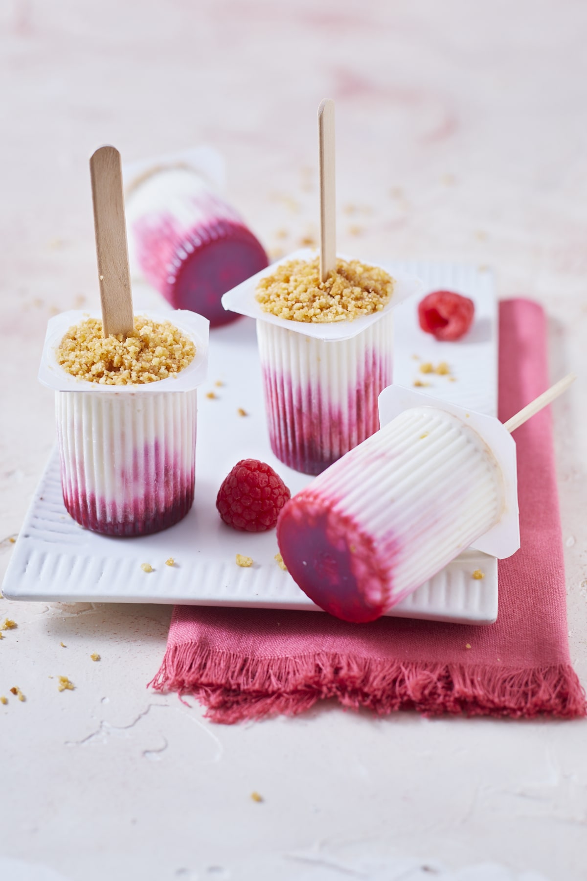 Quatre petits suisses glacés, préparés façon cheesecake à la framboise avec un émietté de galettes bretonnes sur un petit plateau blanc.