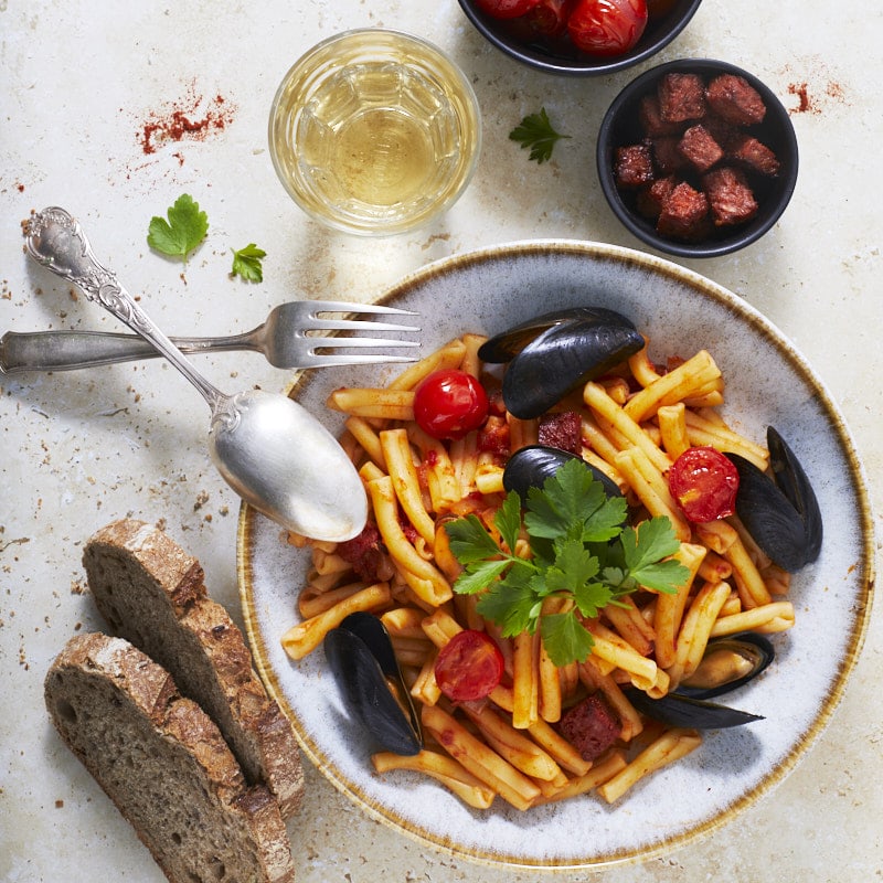 Une assiette de pâtes aux moules à l'espagnole avec des petits bols de dés de chorizo dorés et des petites tomates cerises ainsi qu'un verre de vin blanc.