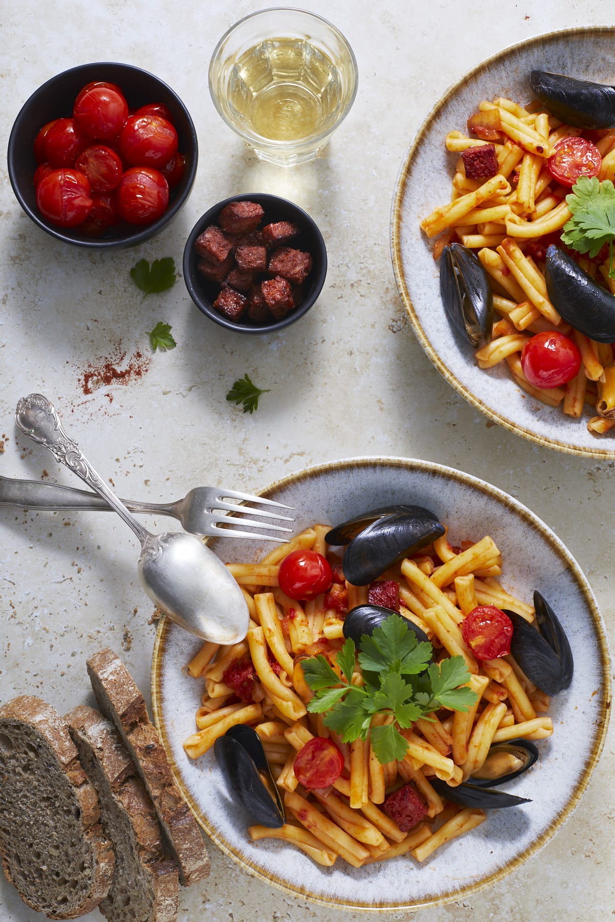 Deux assiettes de pâtes aux moules à l'espagnole accompagnées des petits bols de dés de chorizo et de tomates cerises saisies.