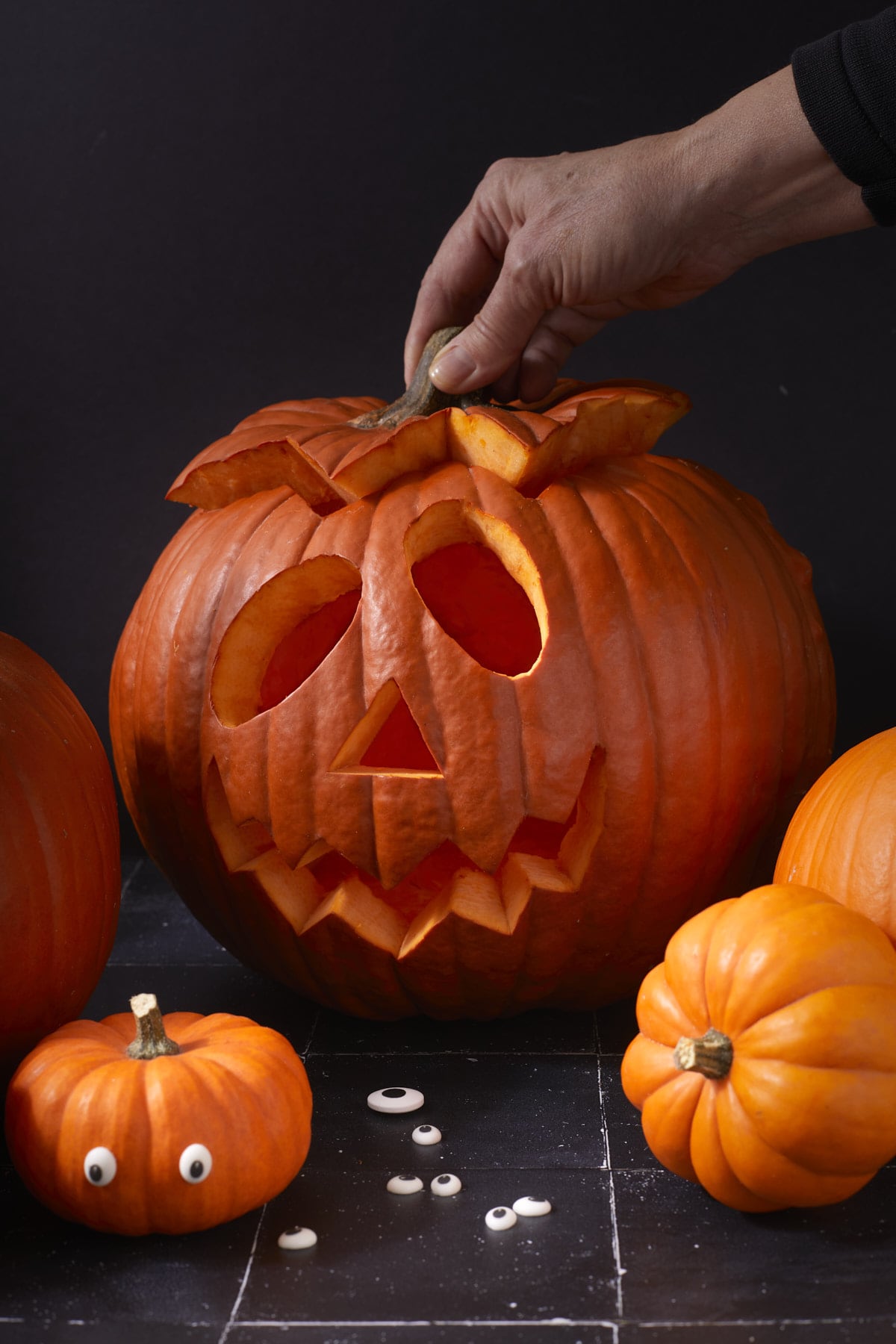 Main soulevant le chapeau d’une lanterne d’Halloween sculptée, entourée de petites courges et d'yeux en sucre